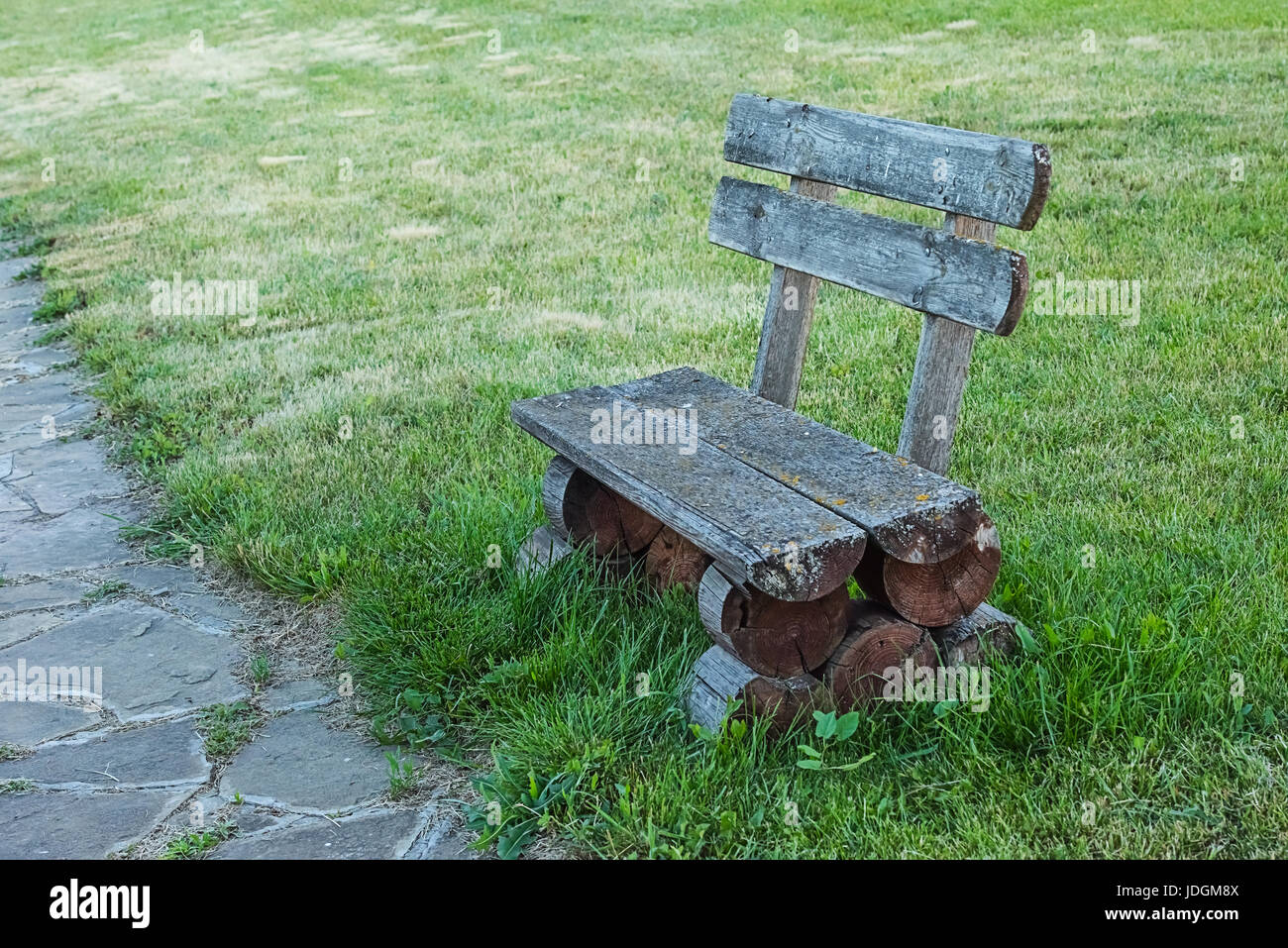 Old handmade wooden bench standing on lawn in the park or garden, from the right of sandstone footpath. Unfocused background. Stock Photo