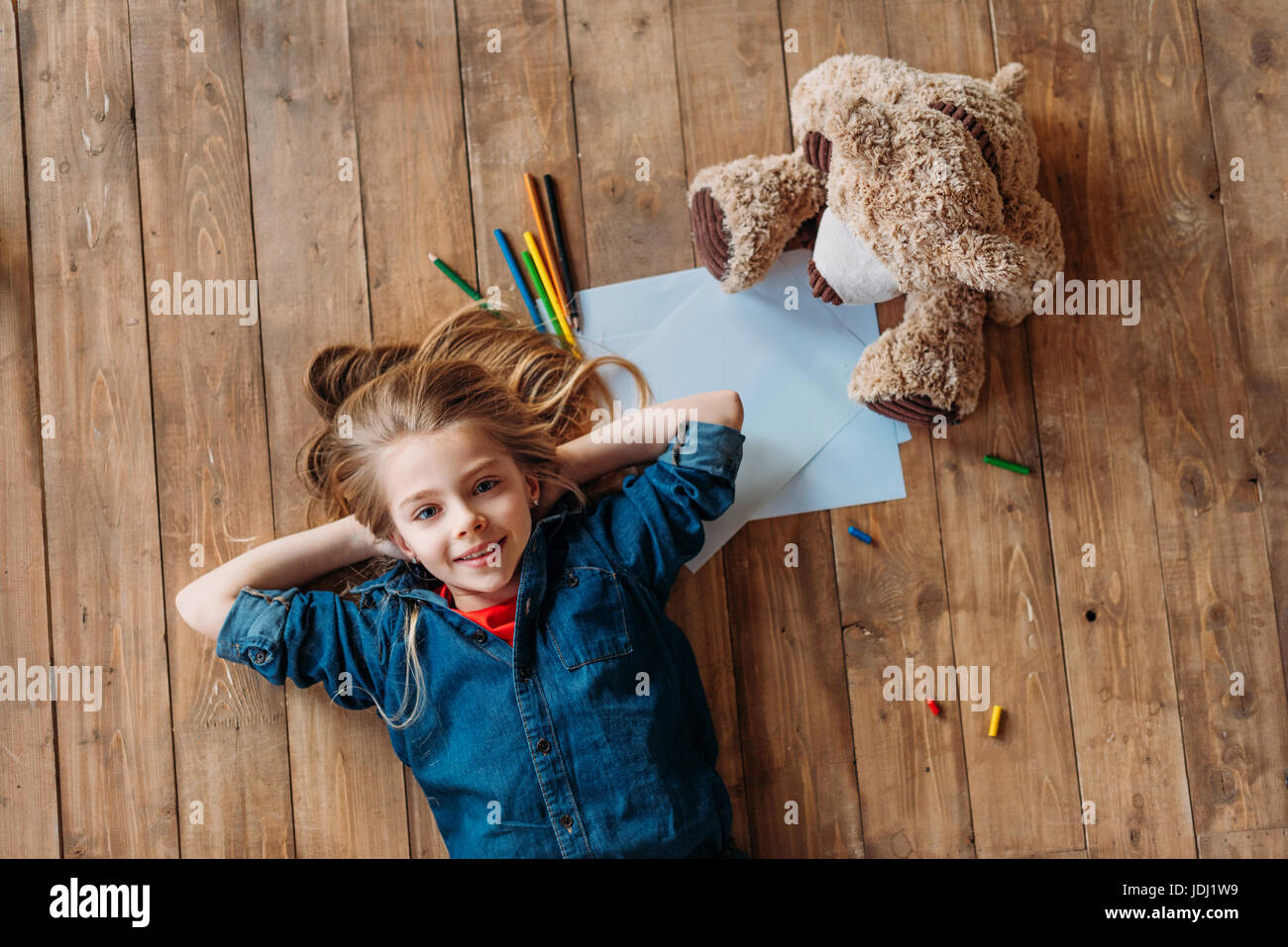 top view of smiling girl lying on floor with teddy bear and pencils ...