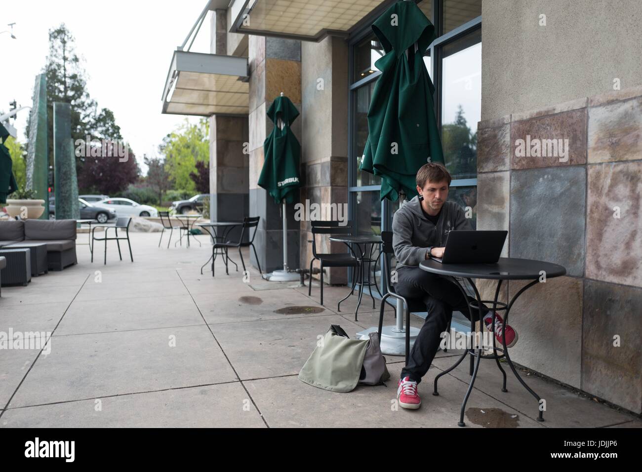 A young male technology worker wearing a hoodie sits outside at a Starbucks and works on a laptop near the Googleplex, headquarters of Google Inc in the Silicon Valley town of Mountain View, California, April 7, 2017. Stock Photo