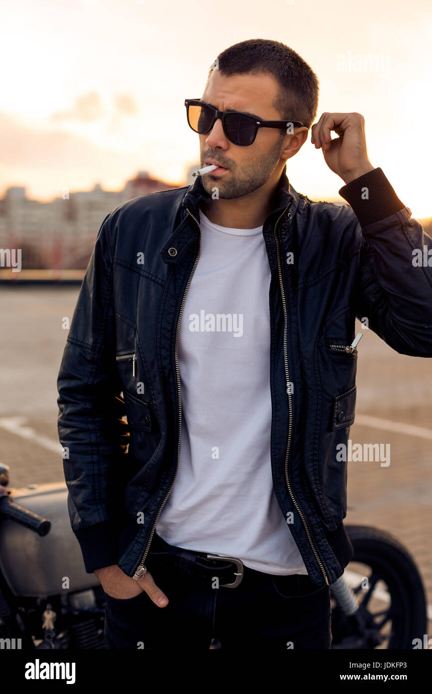 Handsome rider man with beard and mustache in black biker jacket, white t- shirt and sunglasses smoking cigaret near classic style cafe racer  motorbike Stock Photo - Alamy