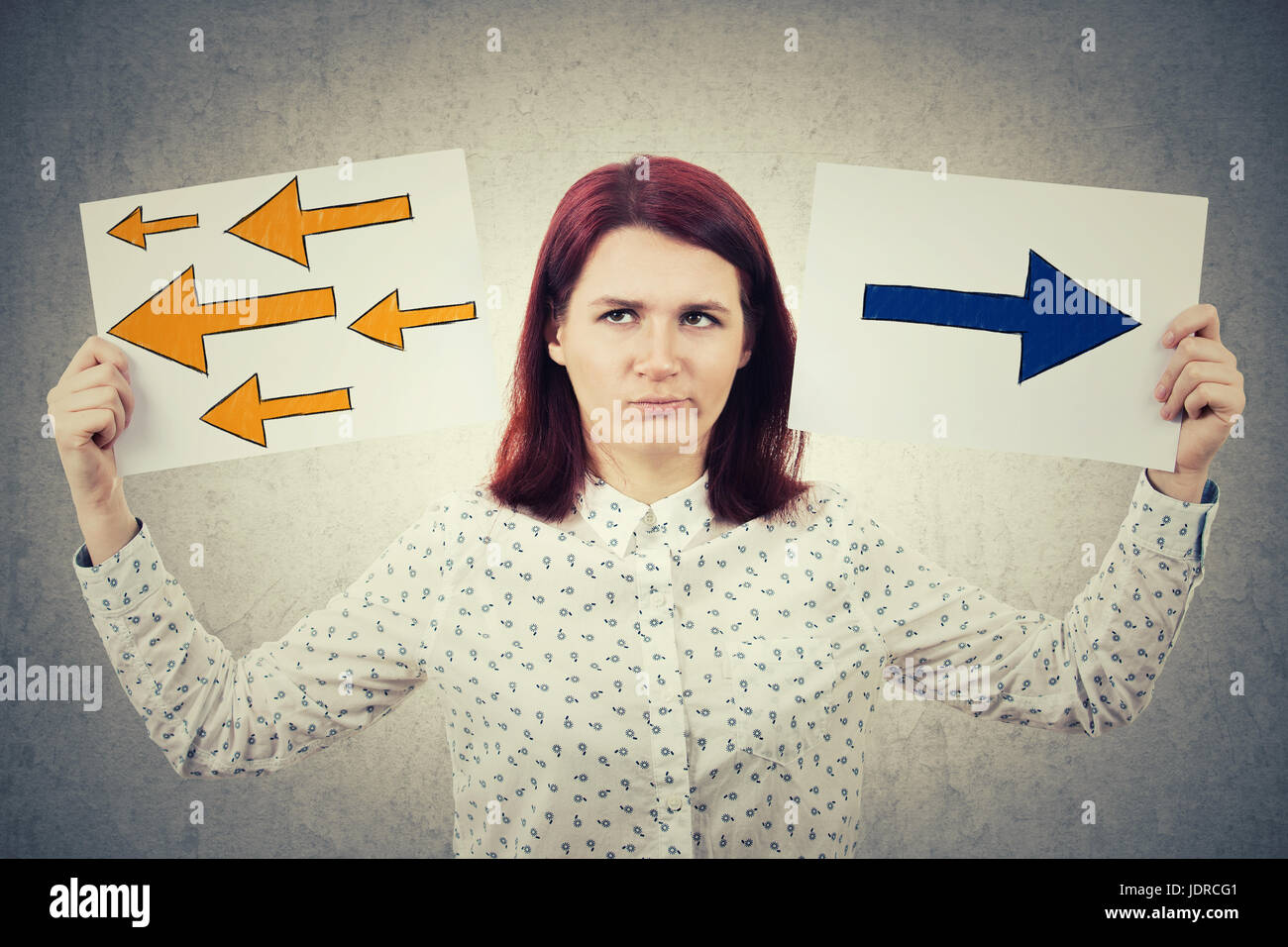 Confused young woman holding two papers in her hands with arrows poinded to the left and right side. Human emotion, face expression life perception co Stock Photo