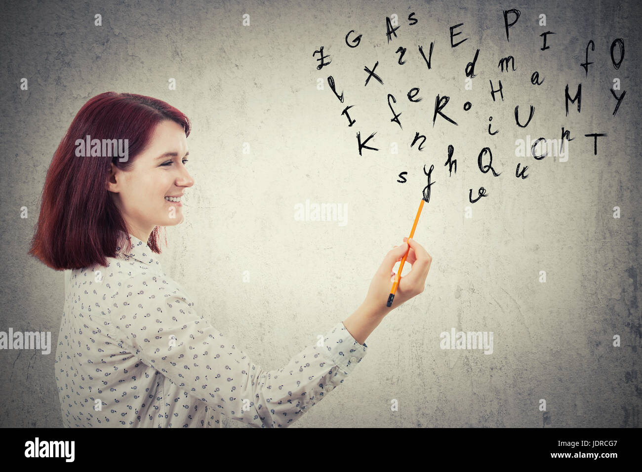 Young businesswoman, holding a pencil in her hand, drawing alphabet letters. Education and communication concept isolated on grey wall background. Stock Photo