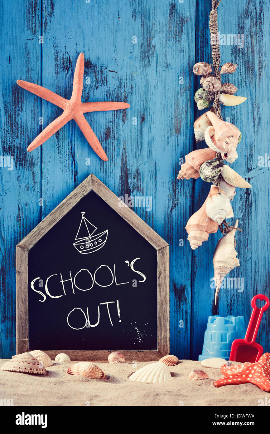 a house-shaped chalkboard with the text schools out surrounded by different beach toys, seashells and starfishes on the sand, against a rustic blue wo Stock Photo