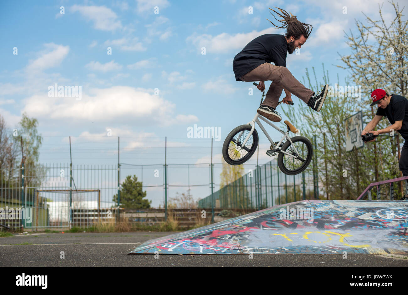 Dreadlock BMX rider performs tailwhip trick in the urban setting of Prague, Czech Republic. Graffiti and blue skies are clear to see. No logos. Stock Photo