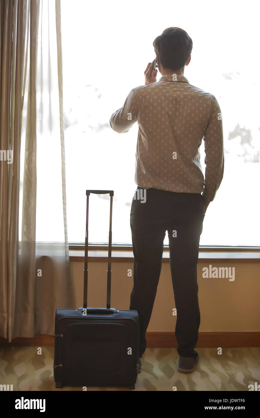 Rear view of man standing near window talking on phone with trolley bag ...