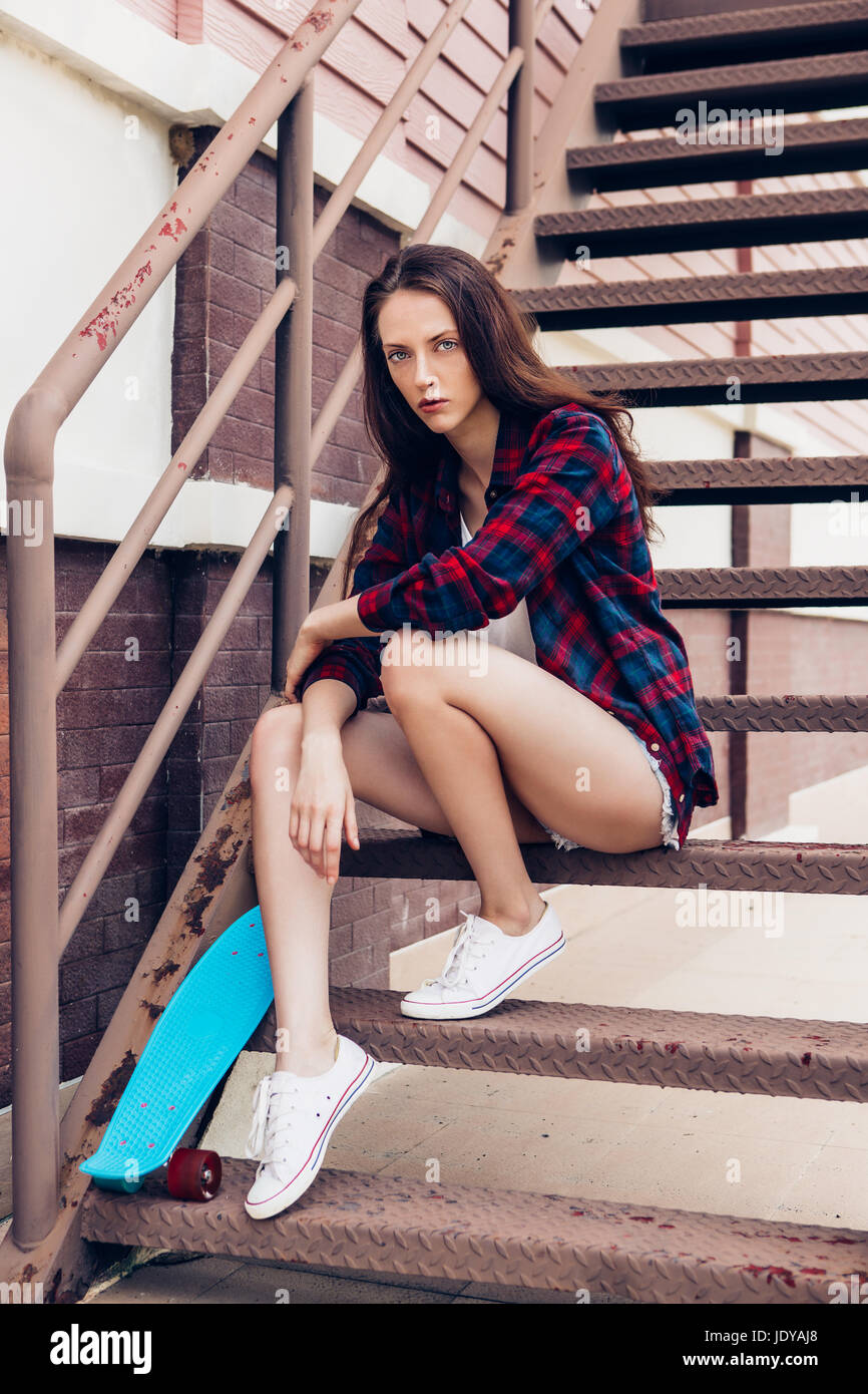 Young beautiful girl in checkered hipster shirt and short jeans shorts and  white sneakers sit on the stairs with her blue mini skateboard longboard. U  Stock Photo - Alamy