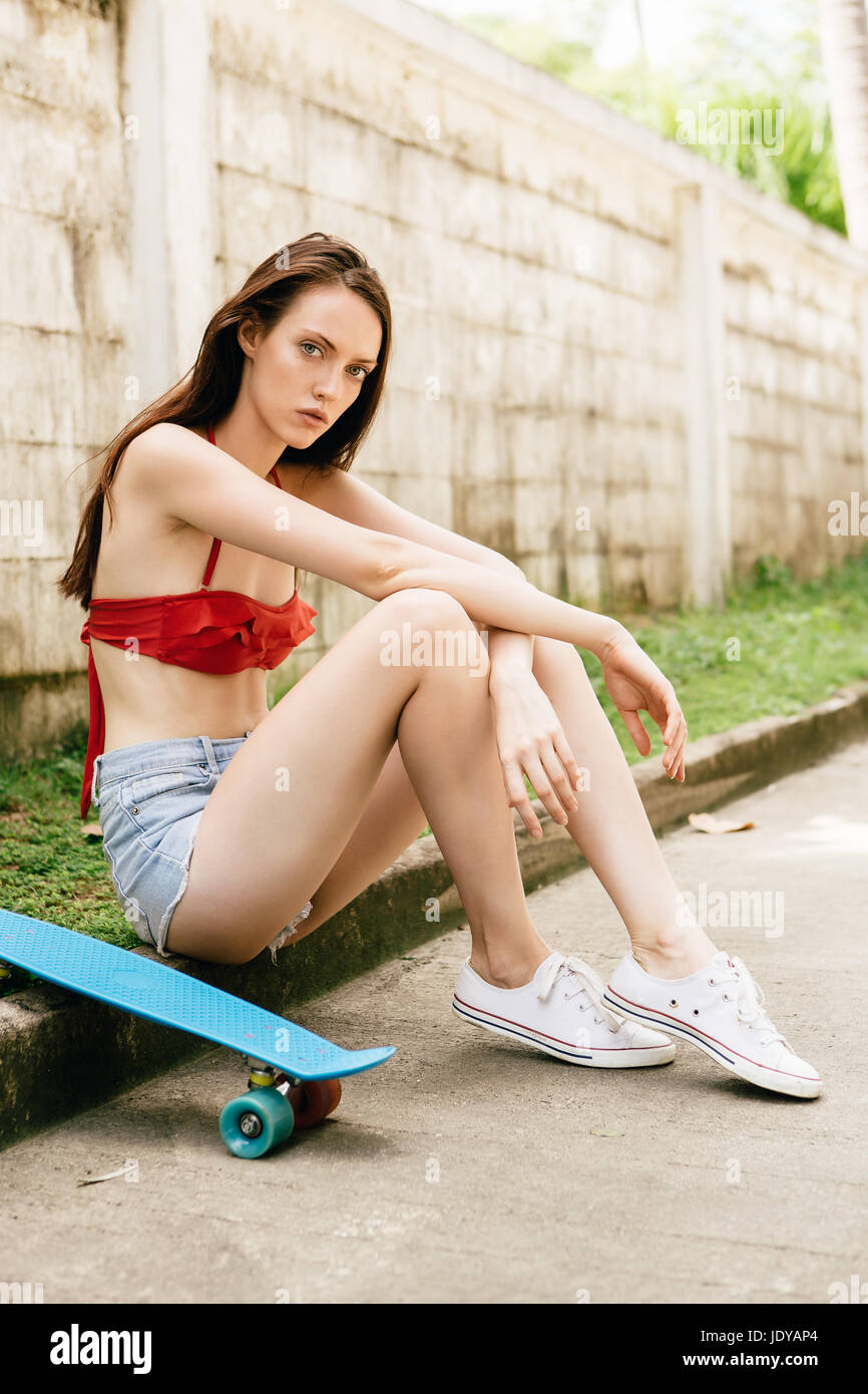 Portrait of pretty beautiful girl in red bra bikini, white sneakers and short denim shorts sit with short skate board. Urban scene, city life. Cute at Stock Photo