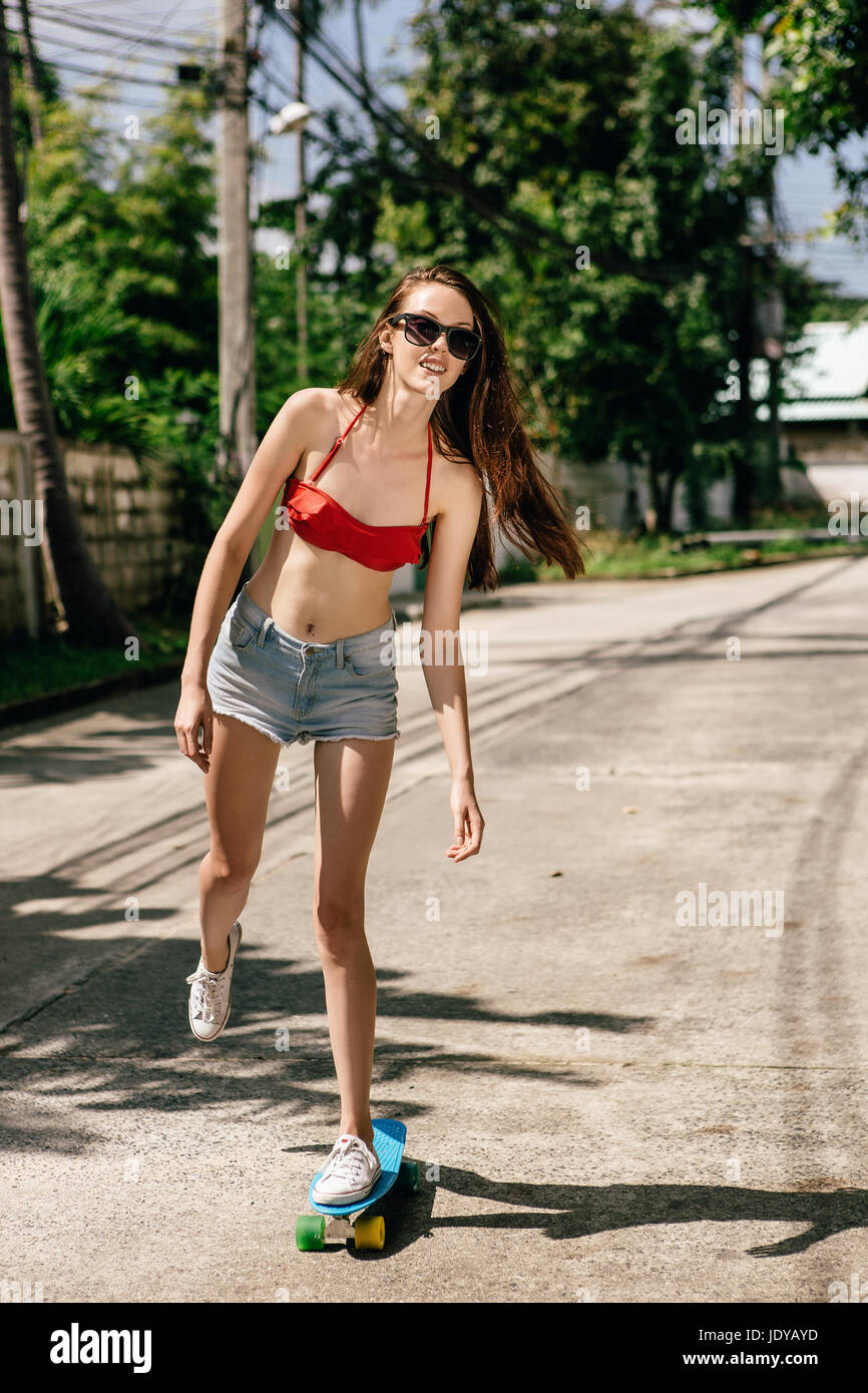 Beautiful girl in red bra bikini, trendy sunglasses, white sneakers and short denim shorts ride blue penny skate board. Urban scene, city life. Cute a Stock Photo