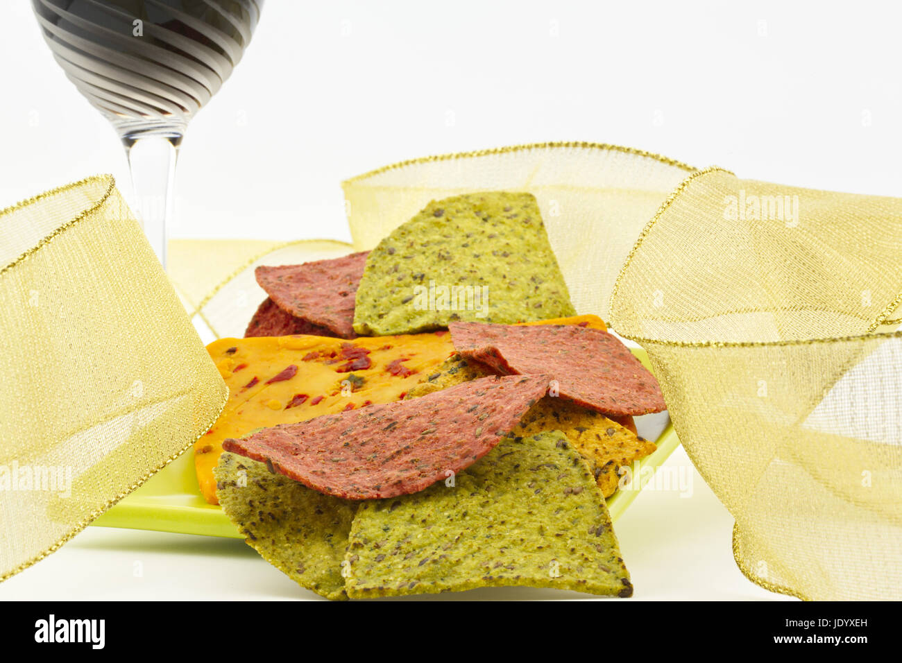 Red wine glass and simple gold ribbon in festive image of party snack foods of cheese with chili and pepper and healthy tortilla chips in green and re Stock Photo