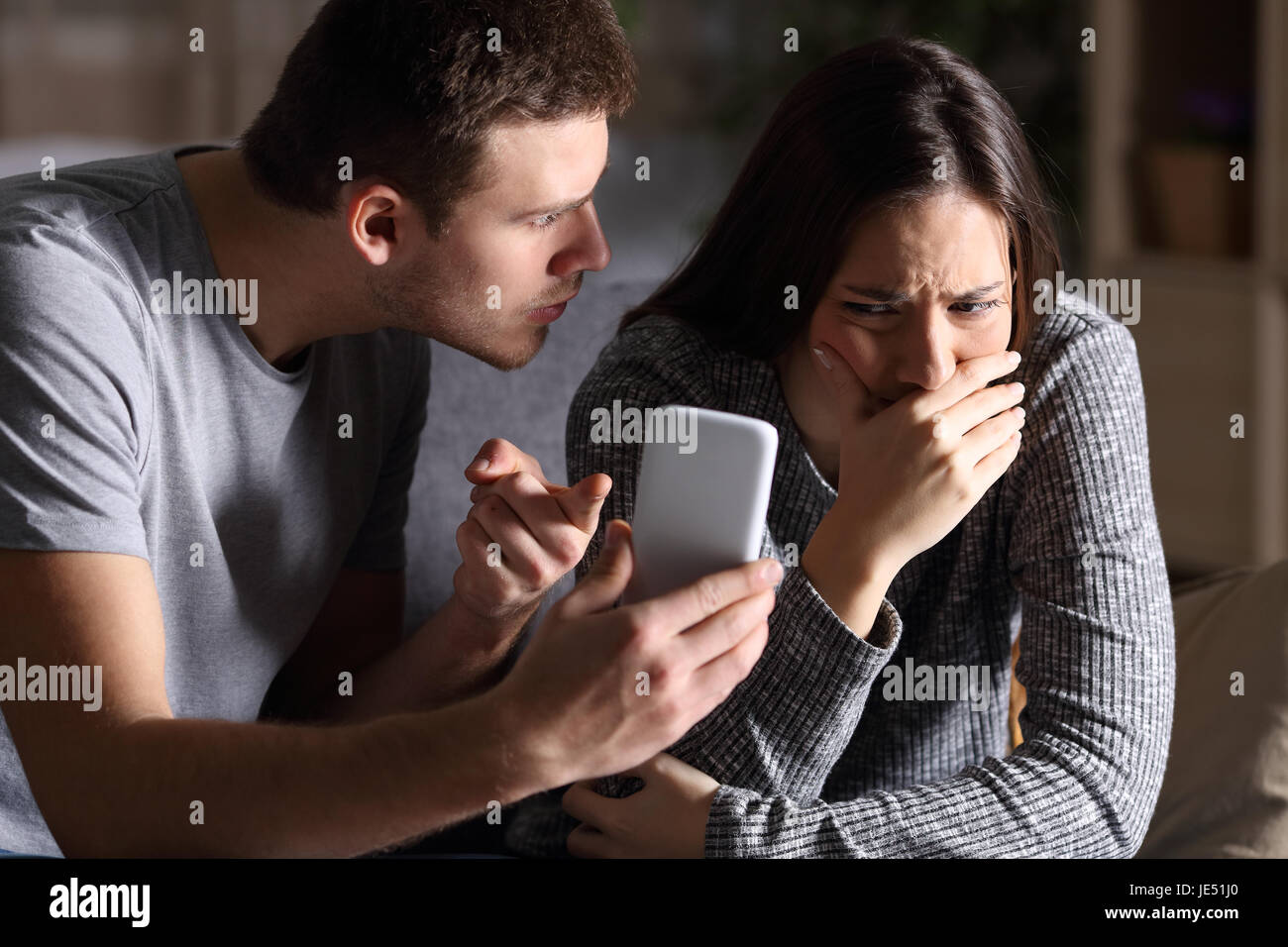 Boyfriend asking for an explanation to his cheater sad girlfriend sitting on a couch in the living room in a house interior with a dark background Stock Photo