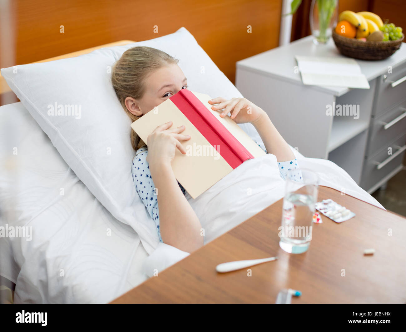 Little girl lying in hospital bed and hiding face with book Stock Photo