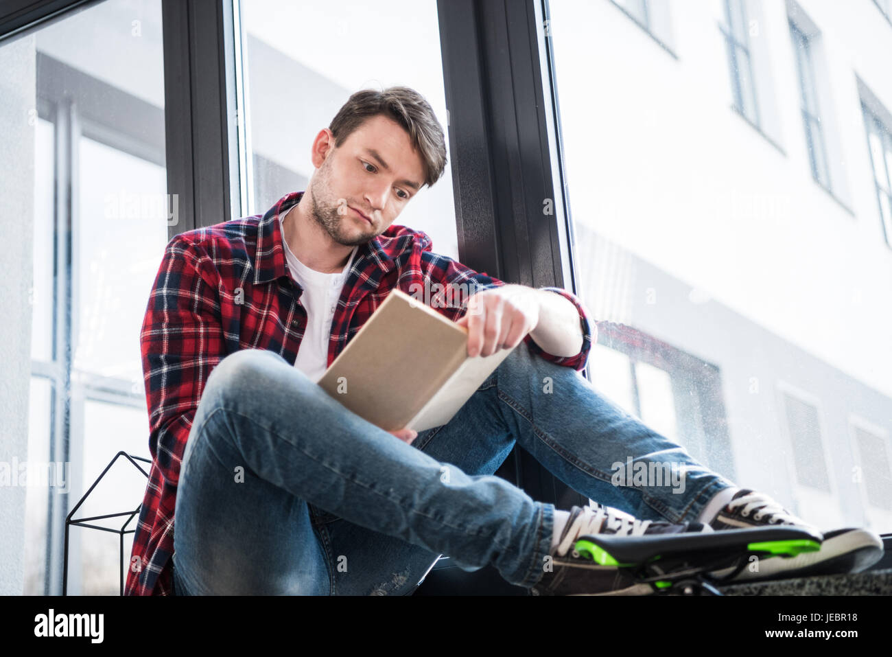concentrated man reading book while sitting on window sill Stock Photo