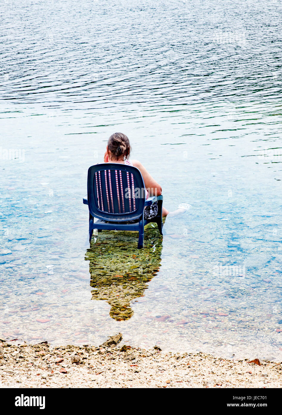 A woman cools off at the beach on a hot day by sitting in a garden chair with her legs in the sea - Croatia Stock Photo