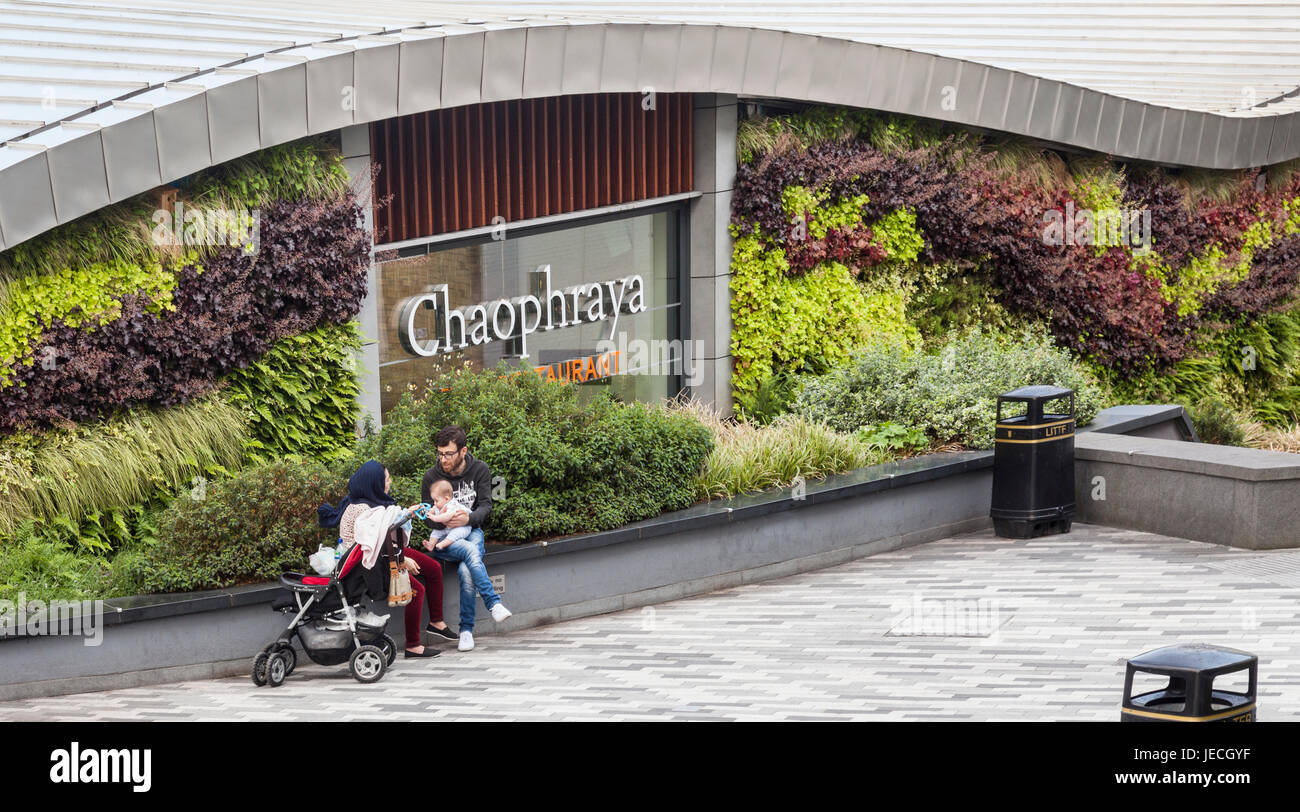 People relaxing on a low wall in front of the living wall at the rear of the Chaophraya Thai Restaurant, in the Bullring, Birmingham, England. Stock Photo