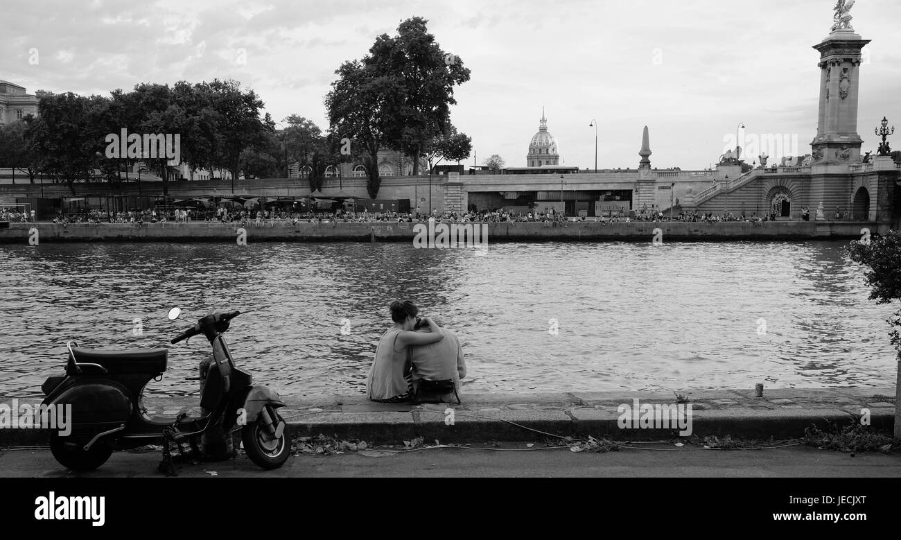 Romantic young couple embrace on the bank of the River Seine in Paris with their scooter nearby Stock Photo