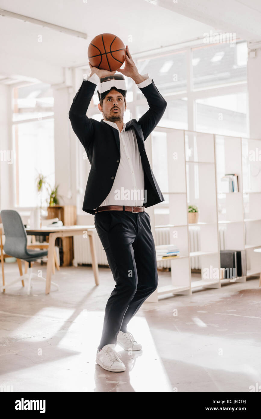 Businessman wearing VR glasses playing basketball in office Stock Photo