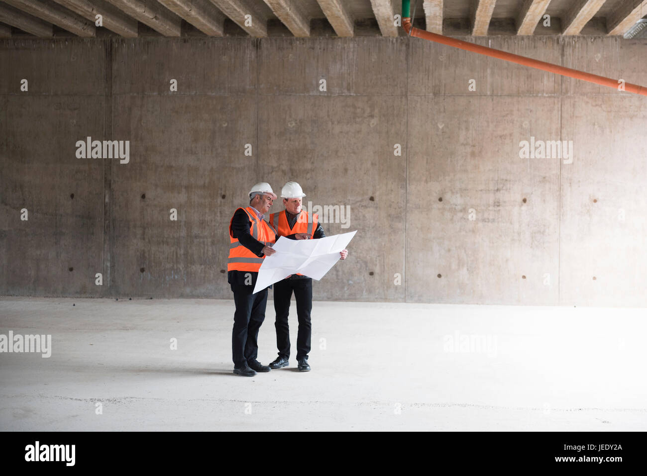 Two men with plan wearing safety vests talking in building under construction Stock Photo