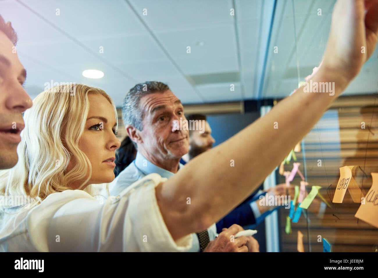 Blond young woman putting sticky note on glass partition in office, workers in background Stock Photo