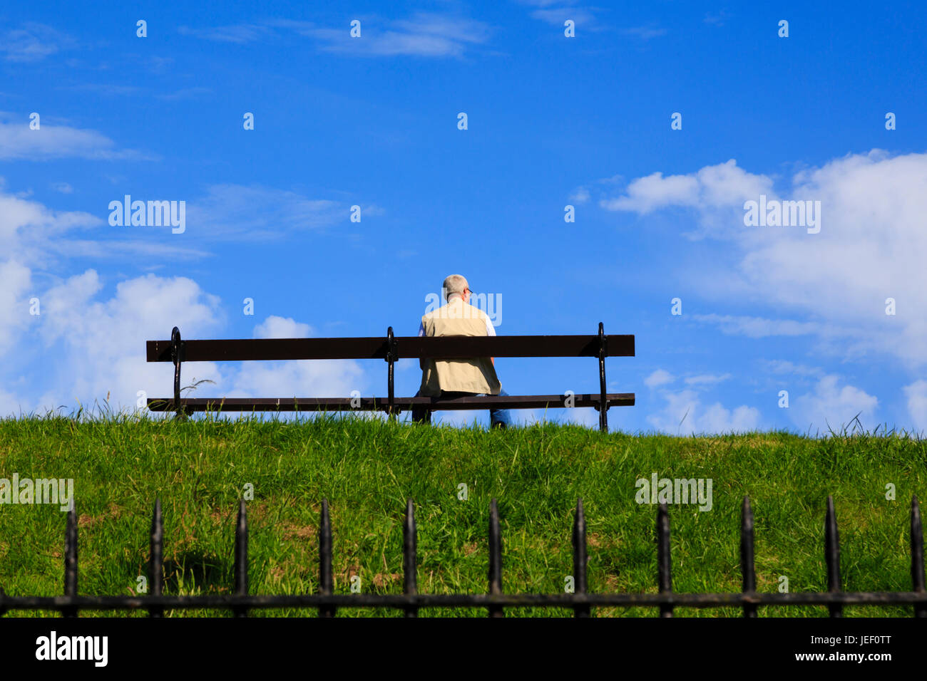 Senior citizen man sitting alone on a bench on a grass bank against a blue sky. On the walls of Berwick upon Tweed. Englands most northerly town. Stock Photo