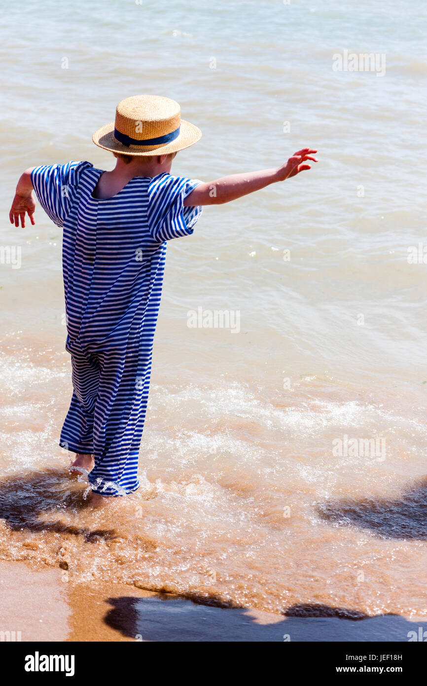 Caucasian child, blonde boy, 4-5 years old, standing in the surf on a beach, wearing straw hat and blue and white Victorian beach costume. Stock Photo