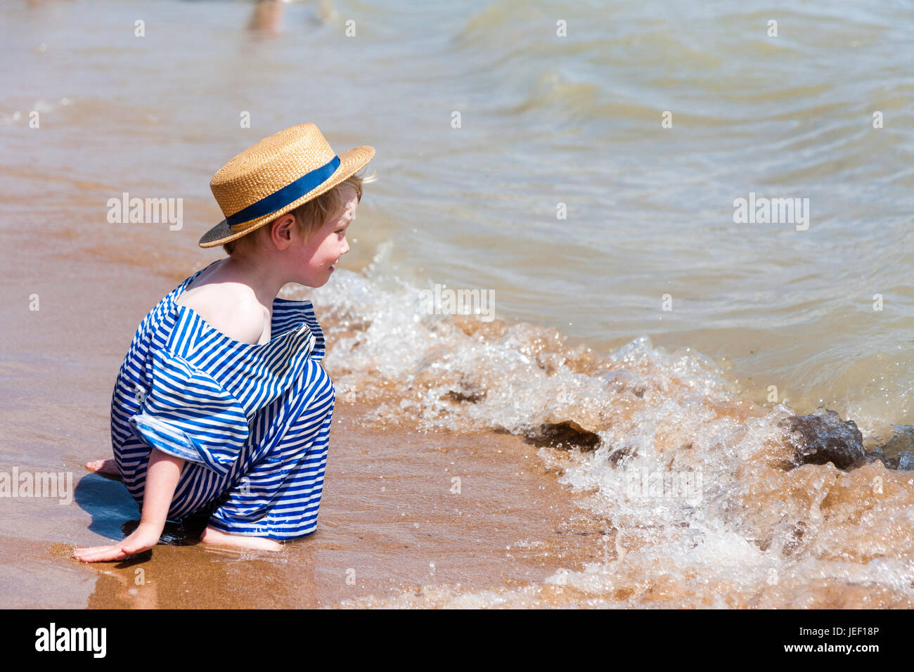Caucasian blonde child, boy, 4-5 years old, kneeling in the surf on a beach, wearing straw hat and blue and white Victorian beach costume. Stock Photo