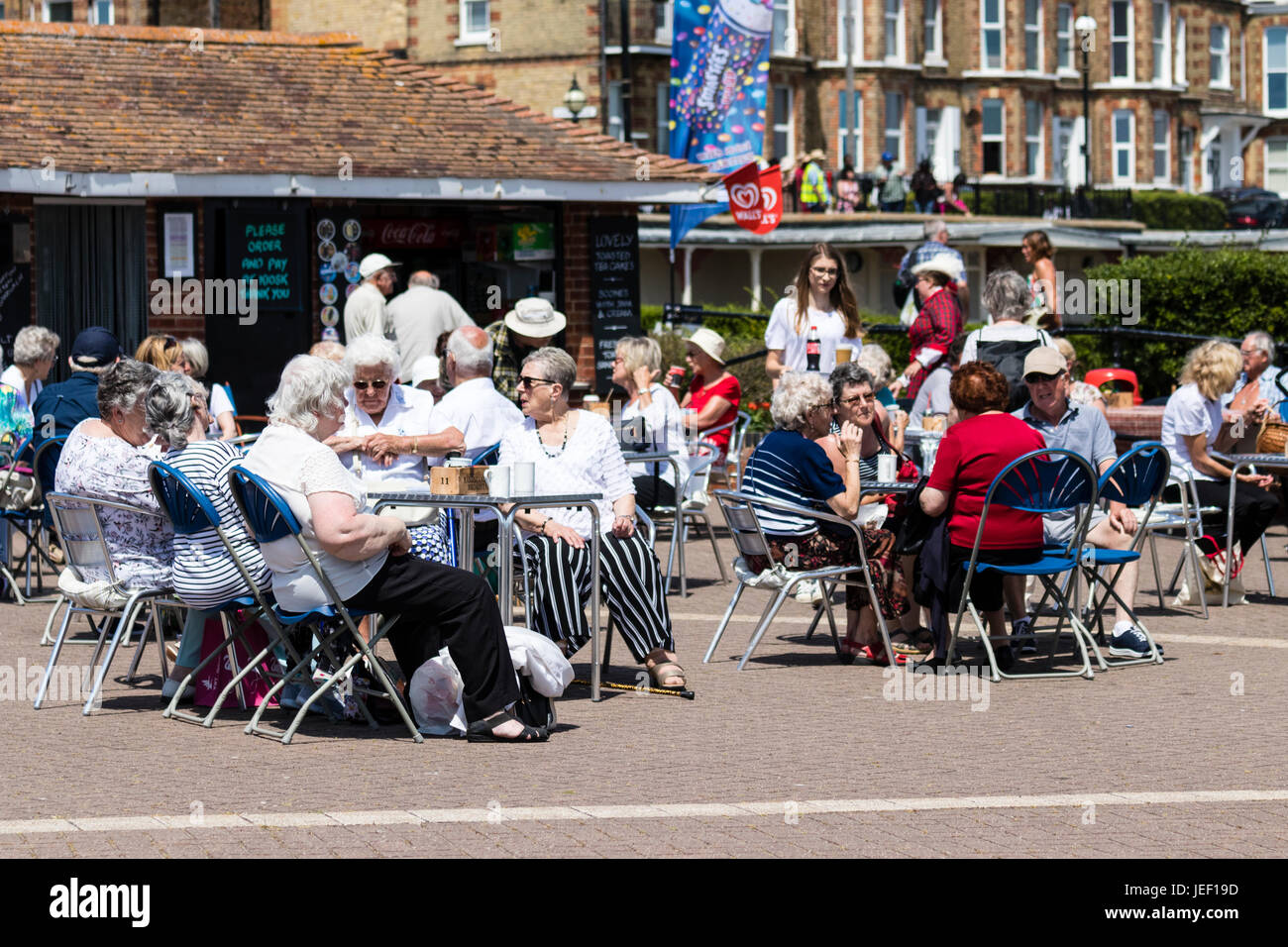 People, mainly seniors, sitting outside on Broadstairs seafront promenade, ay tables by small café kiosk. Bright sunshine, hot weather. Stock Photo