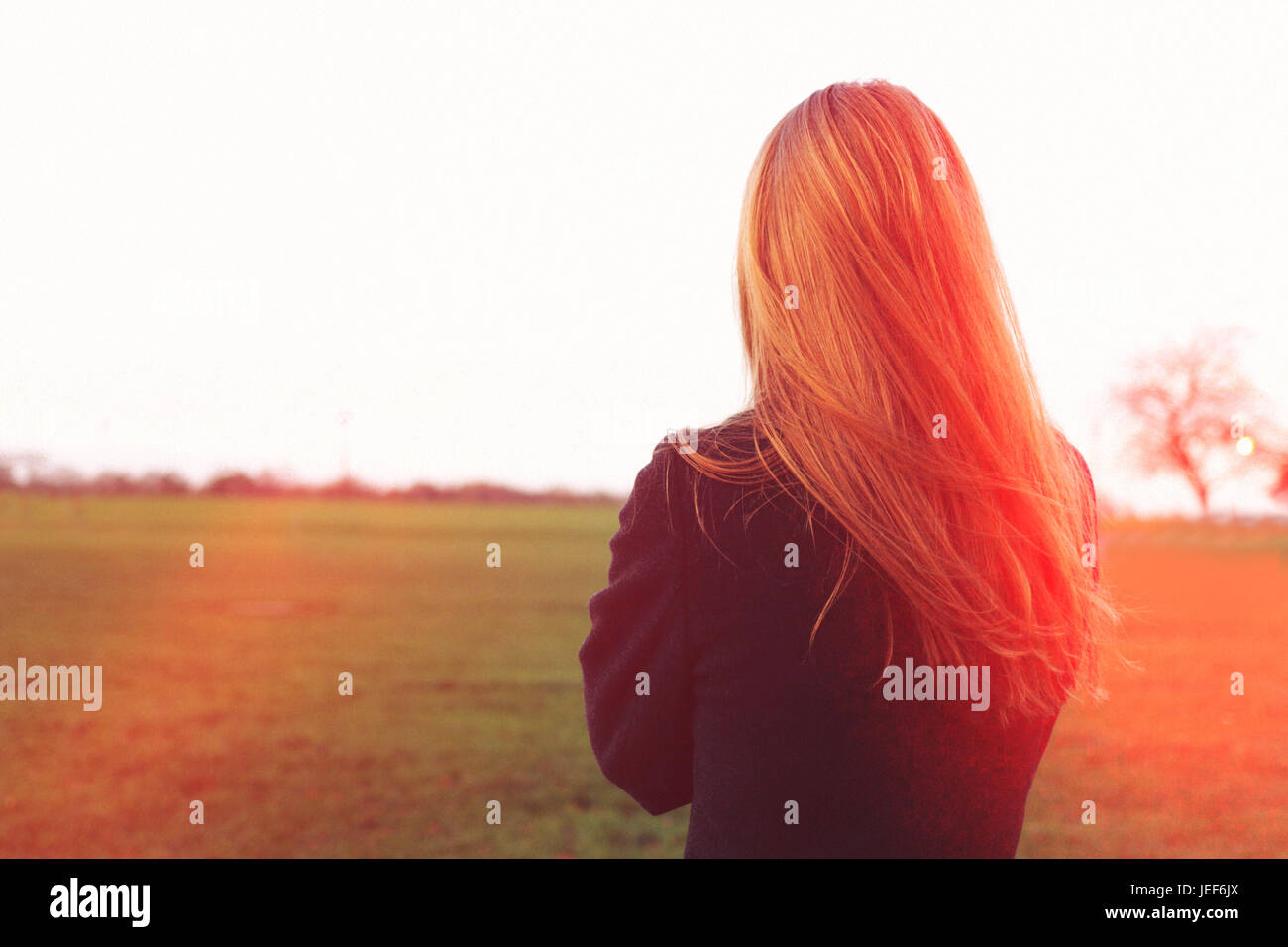 Rear view of a slim young woman standing looking at the horizon in a park. Copy space. Lens flare. She has blonde or red hair and wearing a cot or jam. Stock Photo