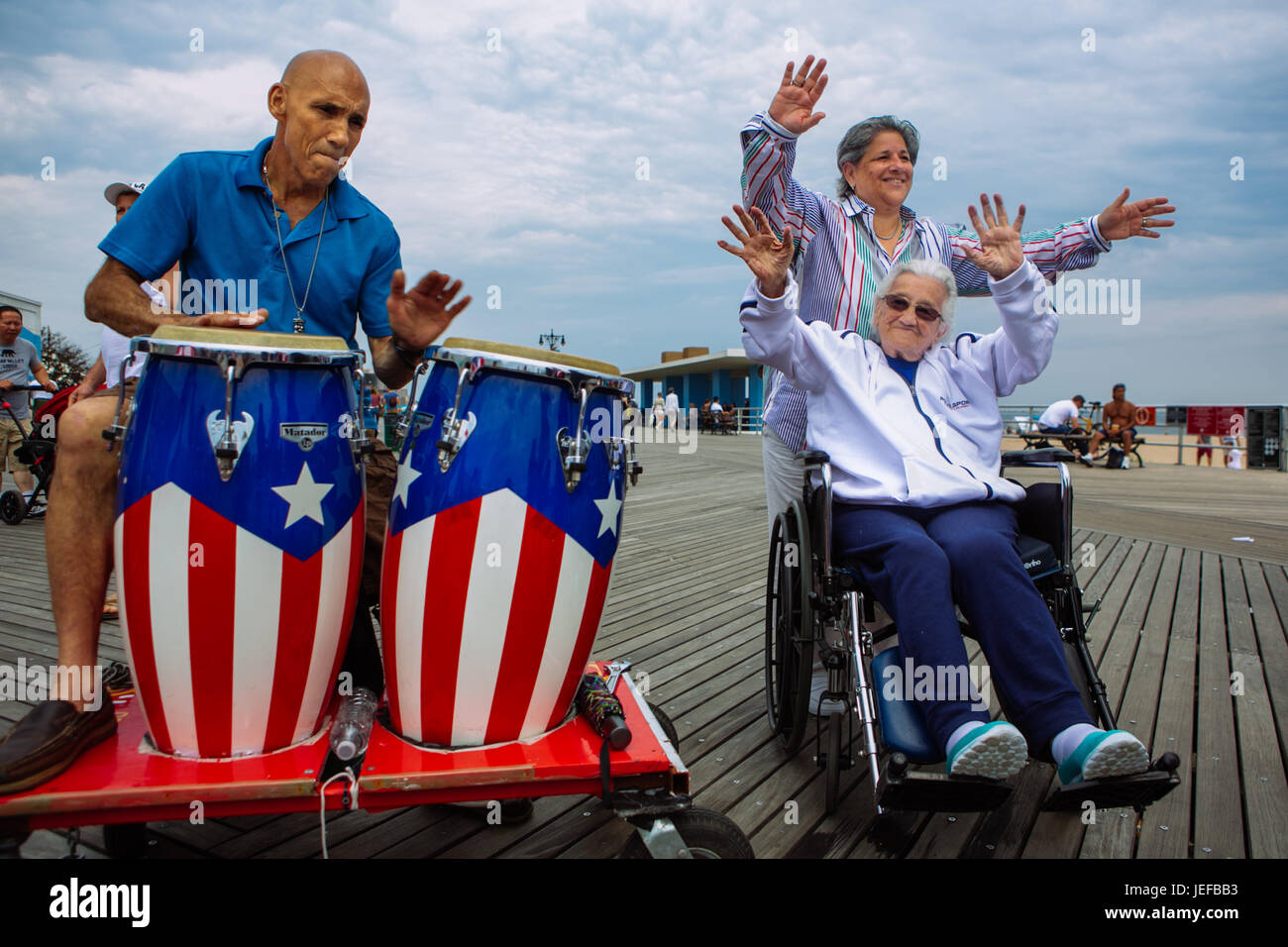 Puerto rico percusion musician playing the drums and an old lady in wheelchair waving arms in Coney Island boardwalk in summer weekend, Brooklyn, USA Stock Photo