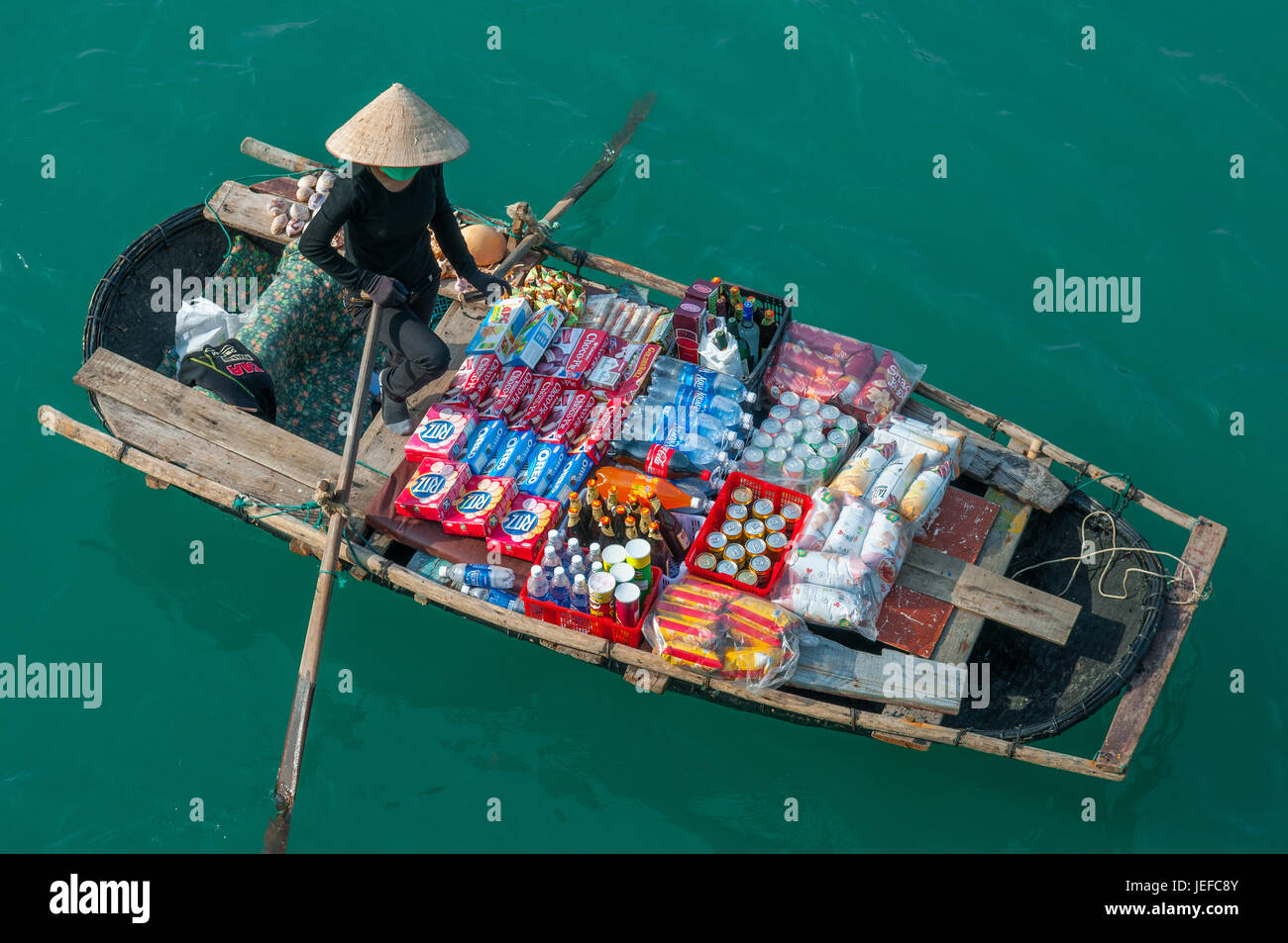 A saleswoman with conical hat in a traditional boat in the emerald waters of Halong Bay selling products to tourist boats, Haiphong, Vietnam. Stock Photo