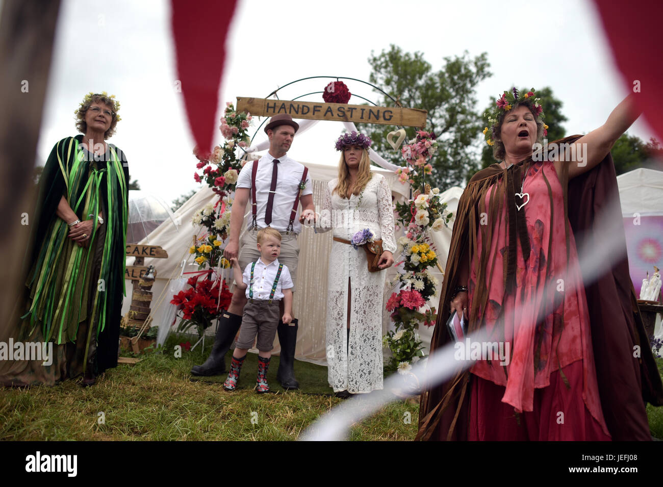 Fi Sexton, 32 and Peter Howe, 38, both from London accompanied by son Thomas Howe, 2 take part in a Handfasting wedding ceremony. The ceremony performed by Glenda proctor (brown) and Jane Tove (green) involved tying their hands together with bonds, Jumping a broom and Eating cake and drinking ale together at Glastonbury Festival, at Worthy Farm in Somerset. Stock Photo