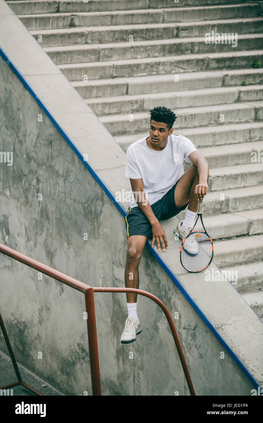 An athlete with a tennis racket sits on a concrete railing of steps ...