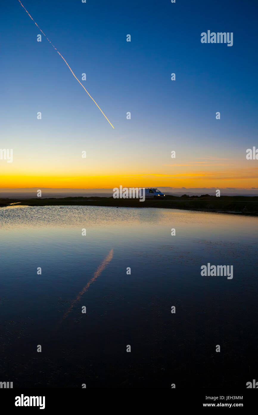aircraft trail at sunset on moors. Vehicle travelling across moors reflected in water at sun. Dartmoor National Park sunset winter, Devon, England, UK Stock Photo