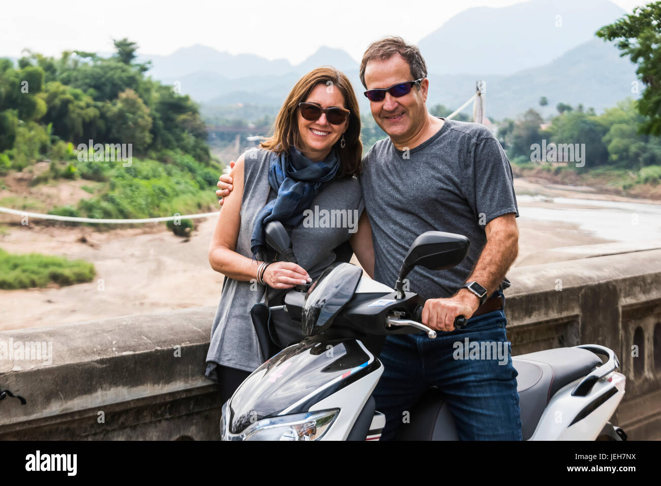 A couple stand posing by a motorcycle with a mountainous landscape in the distance; Luang Prabang, Luang Prabang Province, Laos Stock Photo