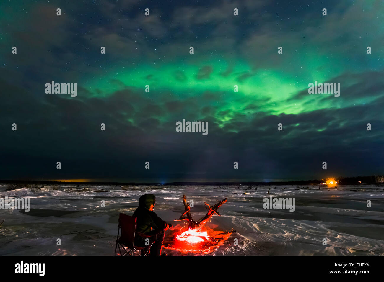 A man warms himself beside a driftwood fire while watching the aurora through clouds on the frozen Delta River, near Delta Junction, Alaska. Stock Photo