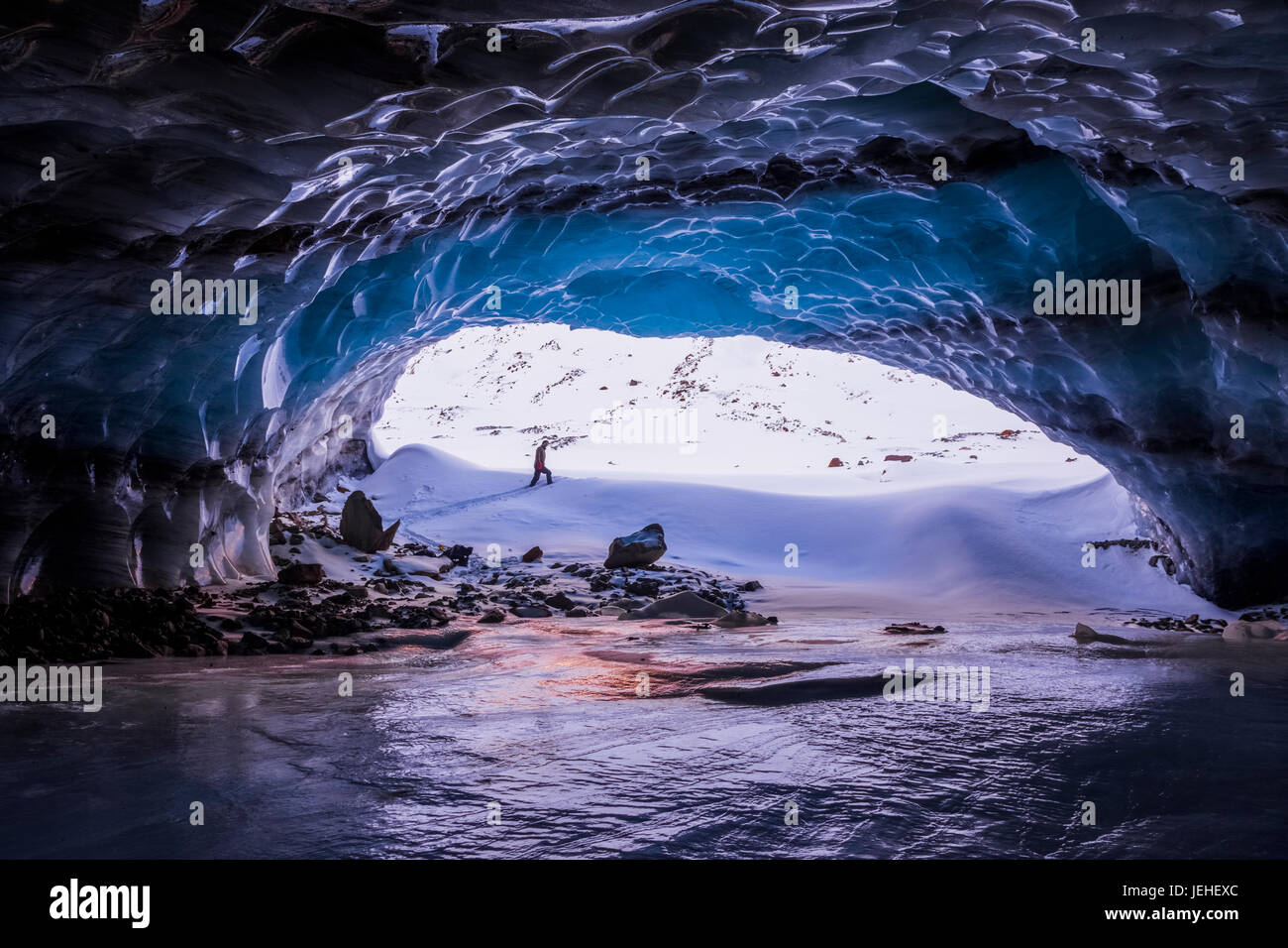A man is framed by the entrance to a large ice cave near the terminus of Augustana Glacier in the Alaska Range in winter Stock Photo