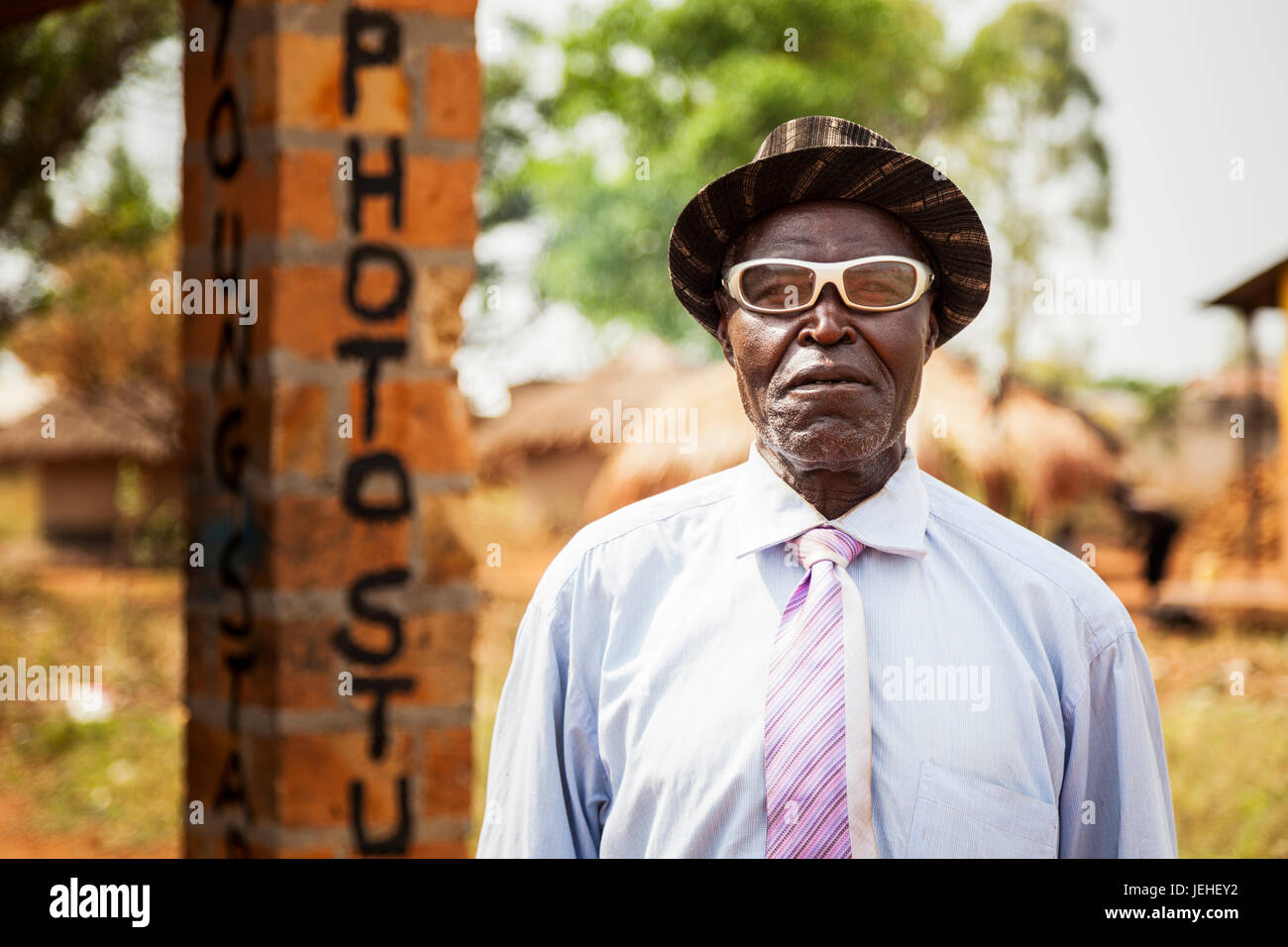 A man with white eyeglasses stands posing outside a photo studio; Uganda Stock Photo