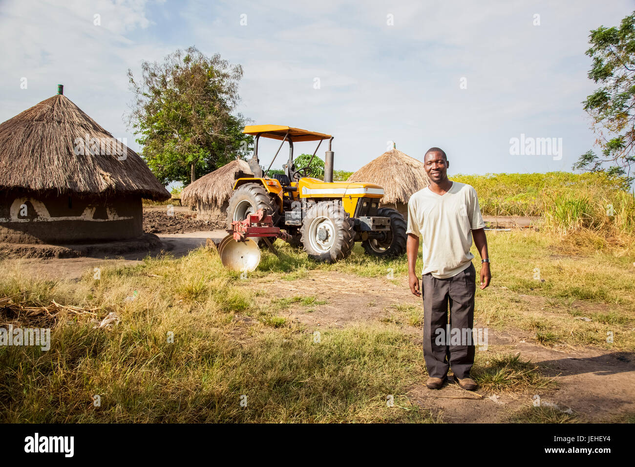 A man stands posing with a tractor and hut in the background; Uganda Stock Photo
