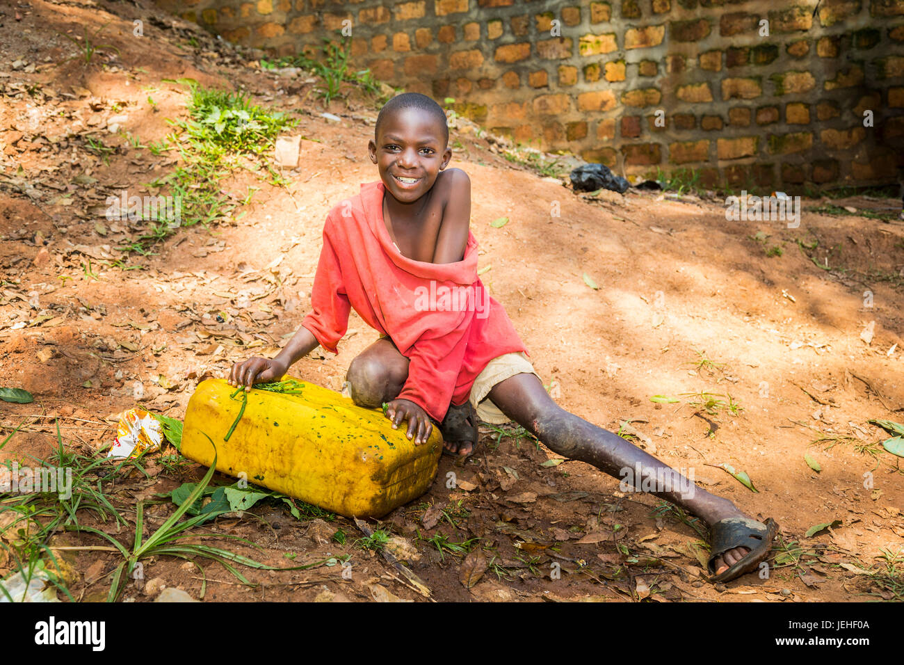 A young boy smiles for the camera while crouching on the ground; Uganda Stock Photo