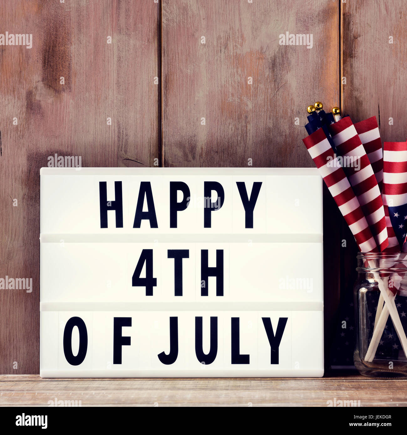 a lightbox with the text happy 4th of july and some american flags on a glass jar, on a rustic wooden background Stock Photo