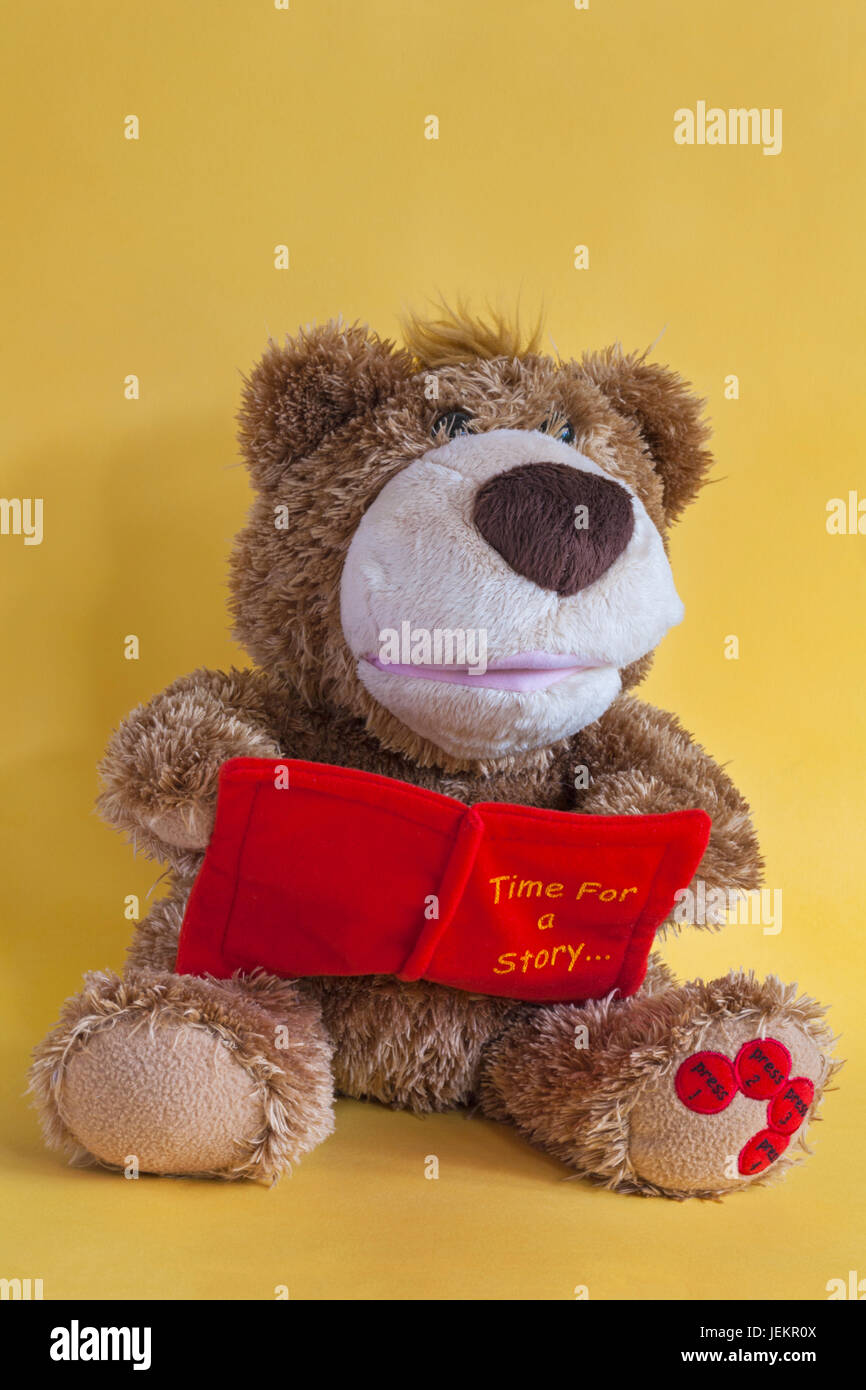 Time for a story - brown coloured teddy bear soft cuddly toy sitting reading story book set against yellow background Stock Photo