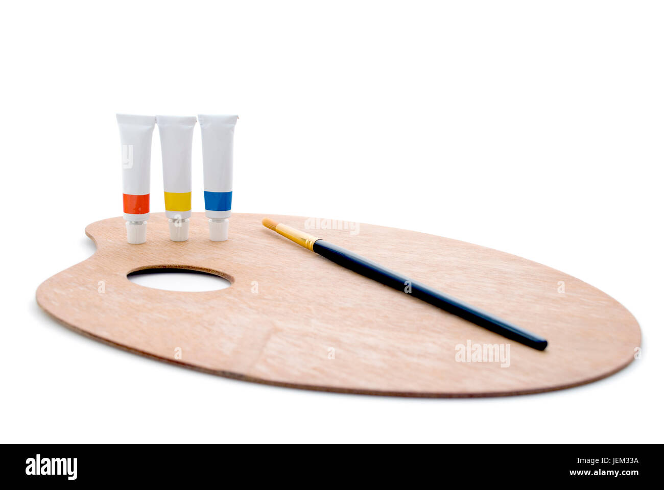 Three tubes of paint (primary colours) and a black/gold paintbrush, standing on a wooden artist's palette, isolated against a white background.  Shado Stock Photo