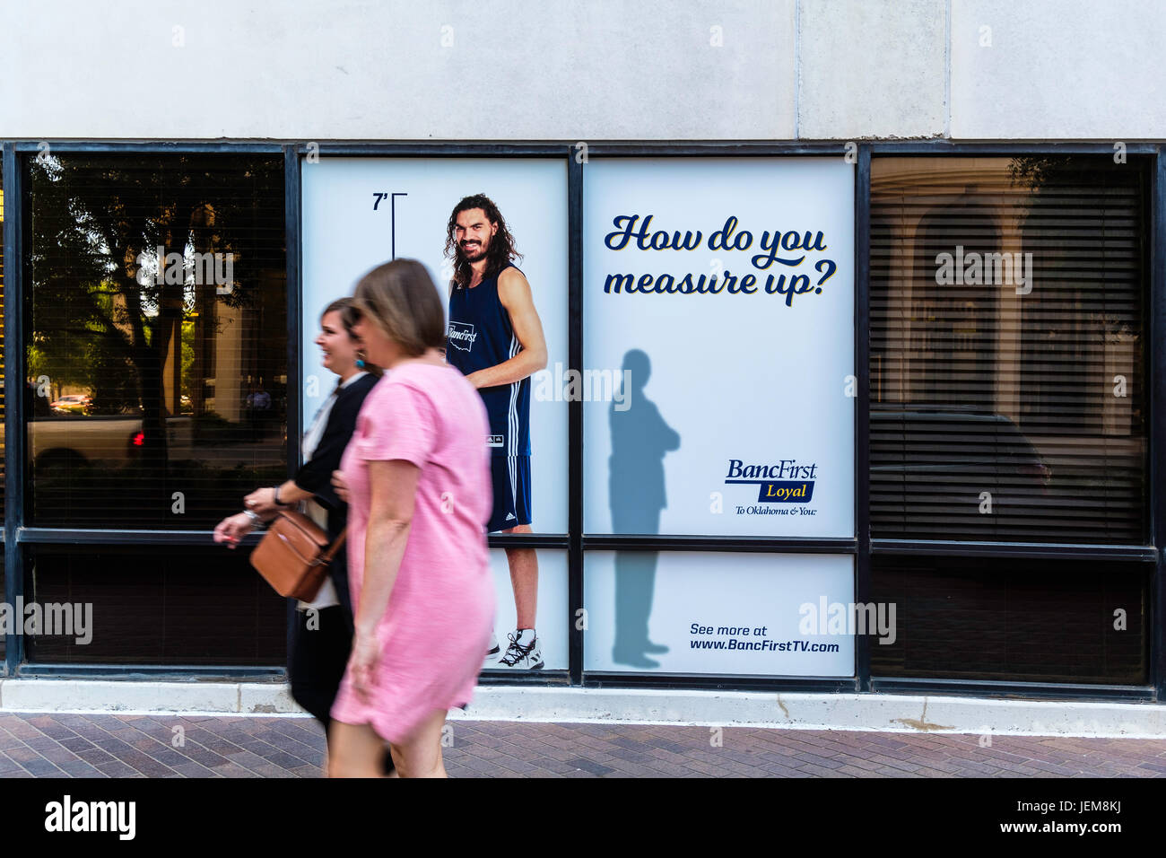 Two women walk past a life sized poster of NBA player Steven Adams in the window of BancFirst in downtown Oklahoma City, Oklahoma, USA. Stock Photo