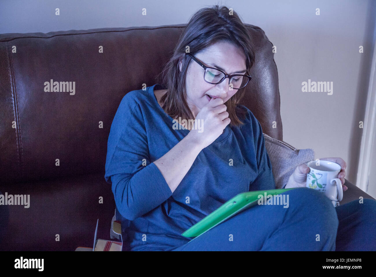 Woman sat on settee using her iPad and holding cup of coffee Stock Photo