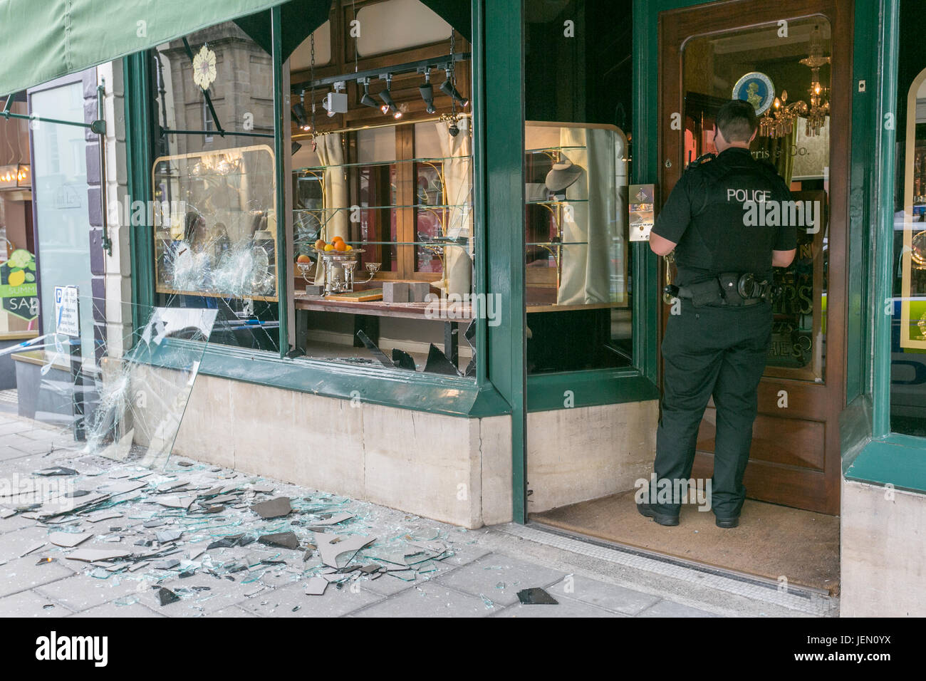 Bristol, UK. 26th June, 2017. A policeman stands next to the smashed jewellery shop windows of Grey-Harris & Co, glass on the pavement. The aftermath of a jewellery 'smash n grab' by 4 armed men who got away on 2 motorbikes in Bristol, UK. Credit: Christian Lathom-Sharp/Alamy Live News. Stock Photo