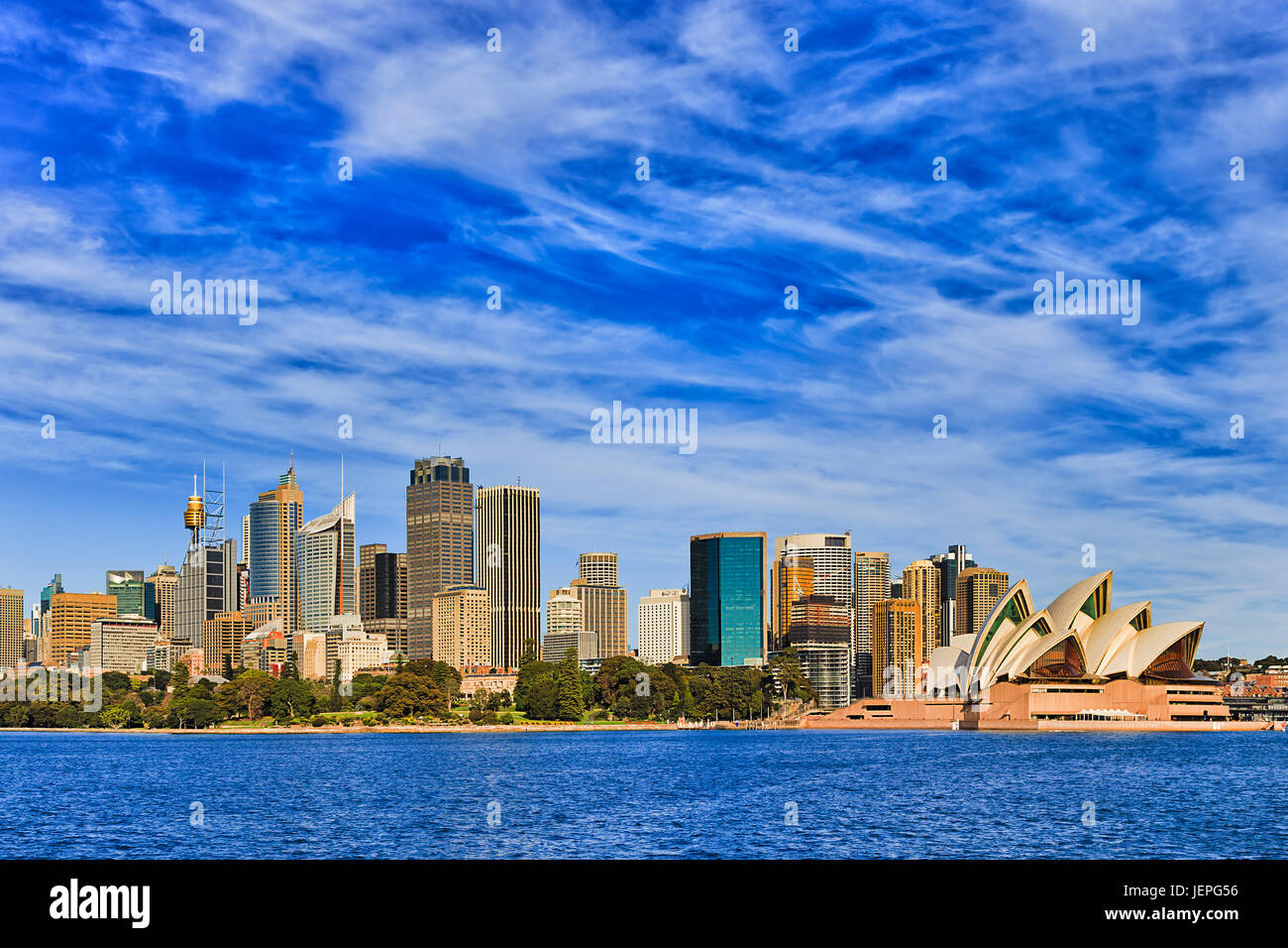 Cityscape of Sydney CBD across Royal Botanic gardens as seen from Sydney harbour ferry with blue water and sky. High-rise towers lit by bright warm su Stock Photo