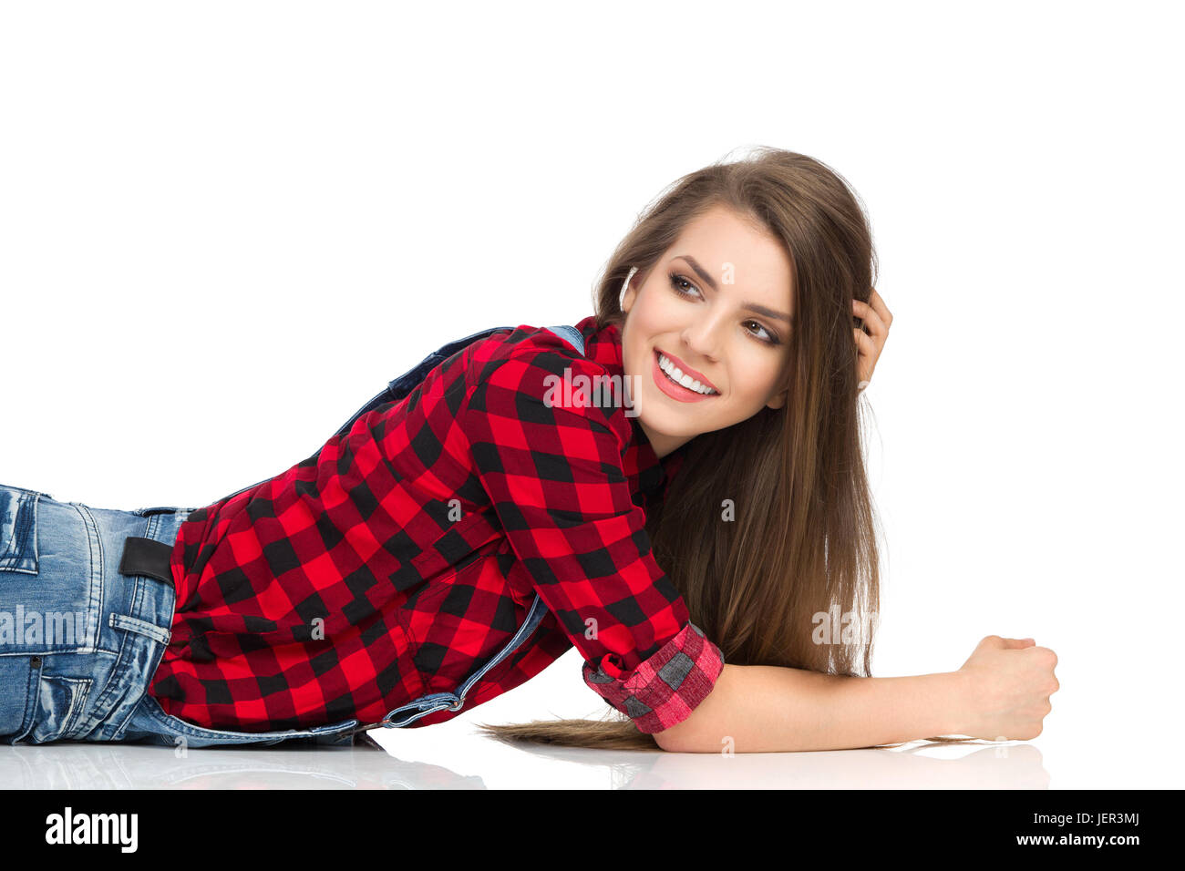 Smiling young woman in red lumberjack shirt and jeans  lying down on a floor and looking away. Waist up studio shot isolated on white. Stock Photo