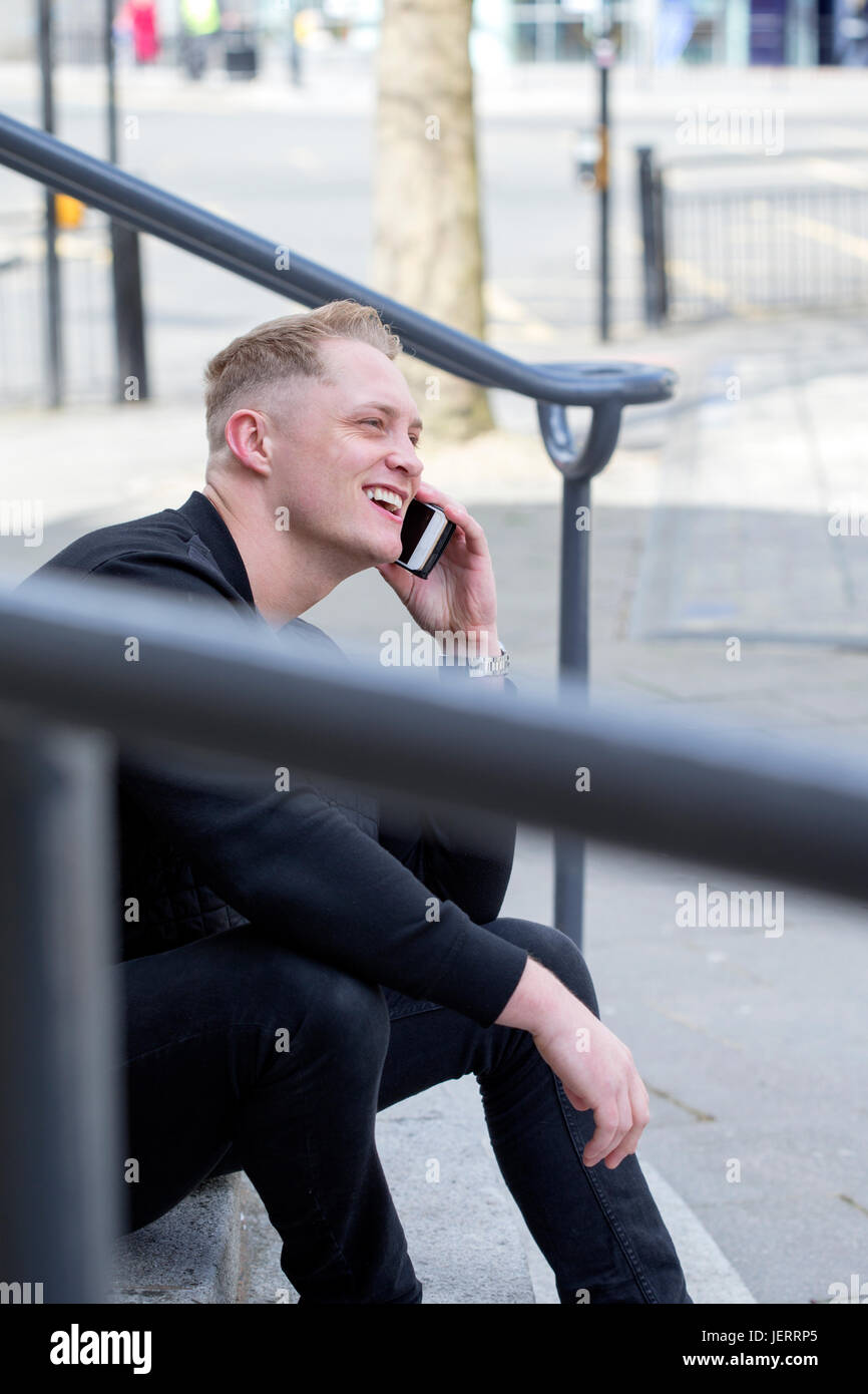 Young man sitting on some steps in the city. He is using his smartphone to have a phonecall. Stock Photo