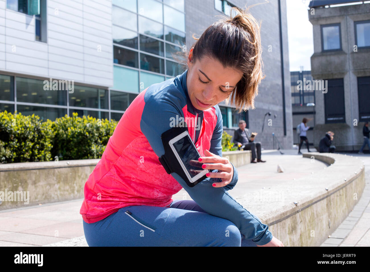 Woman taking a break from her run in the city. She is sitting on a wall and is using the smartphone that is in a strap round her arm. Stock Photo