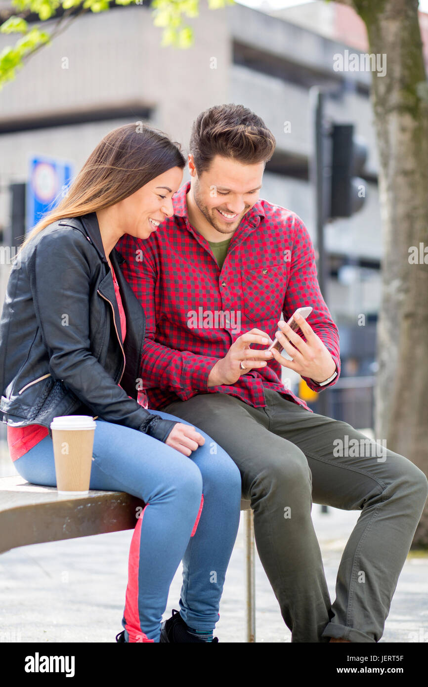 Young couple sitting in the city. The man is holding a smartphone, which they are both looking at. Stock Photo