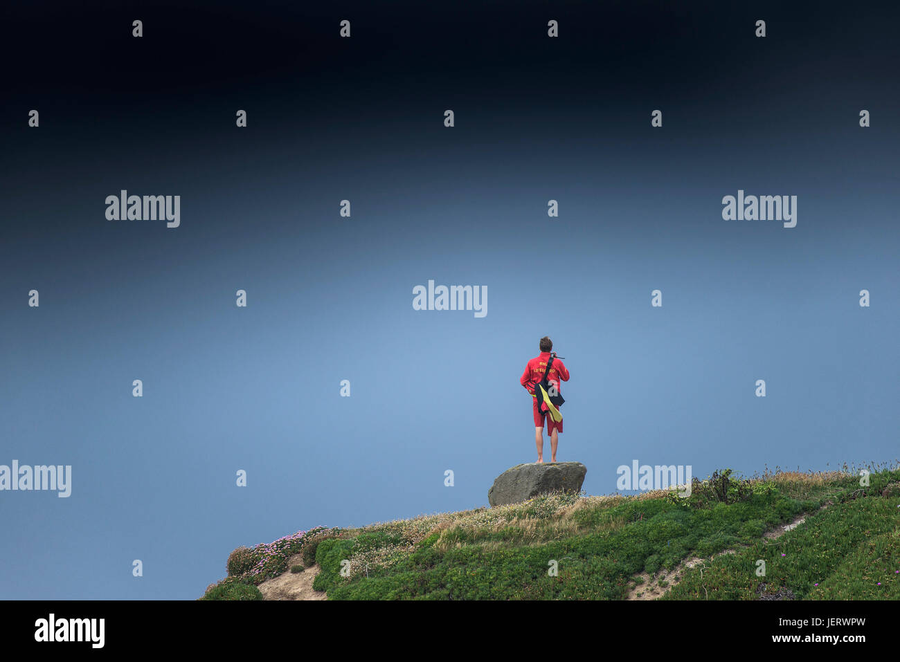 A RNLI Lifeguard standing on a rock using a personal radio as storm clouds gather. Stock Photo
